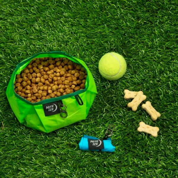 Bolsa de comida para perros con croquetas, pelota de tenis y huesos de juguete en césped artificial.